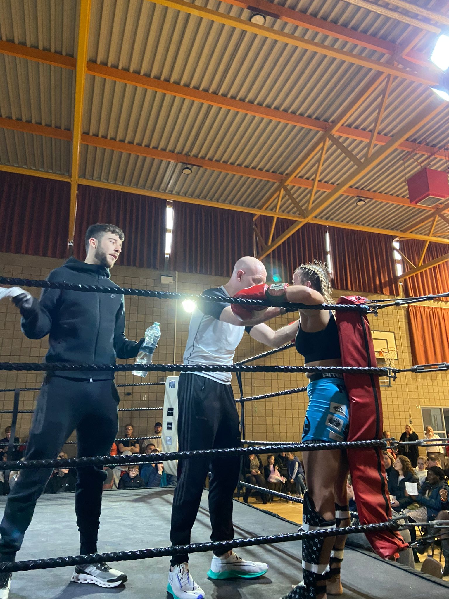 Amber Lennon leaning in the corner of a boxing ring between rounds, getting advice from Phil Whyte and being fanned by Jordan Dempsey